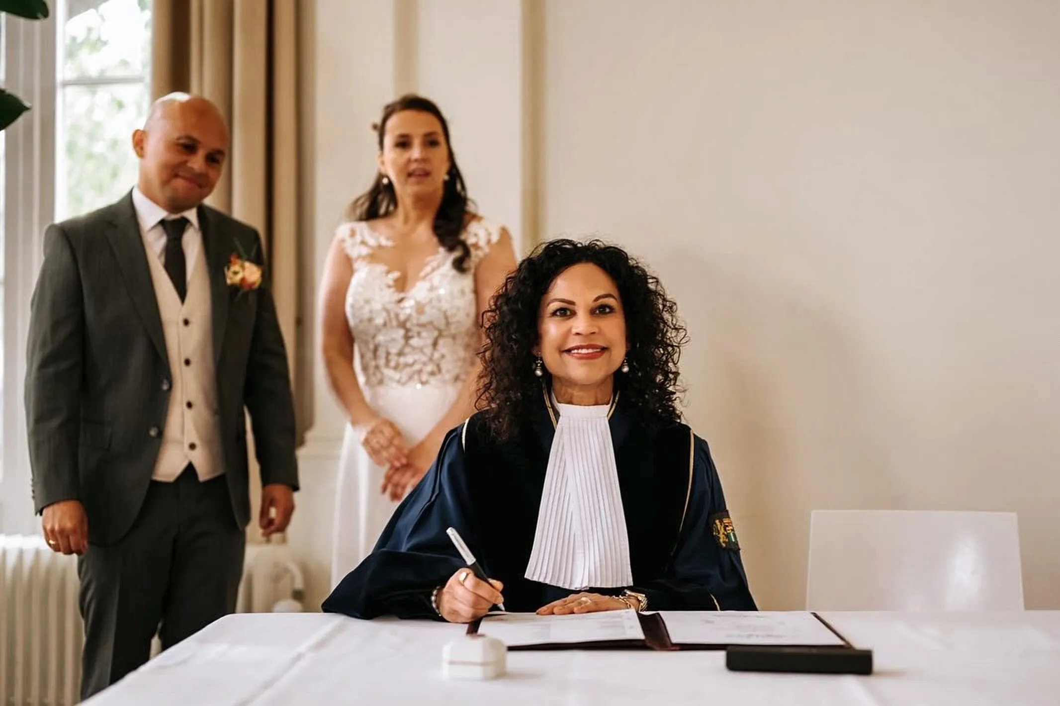 Angela tekent de huwelijksakte terwijl het bruidspaar achter haar toekijkt